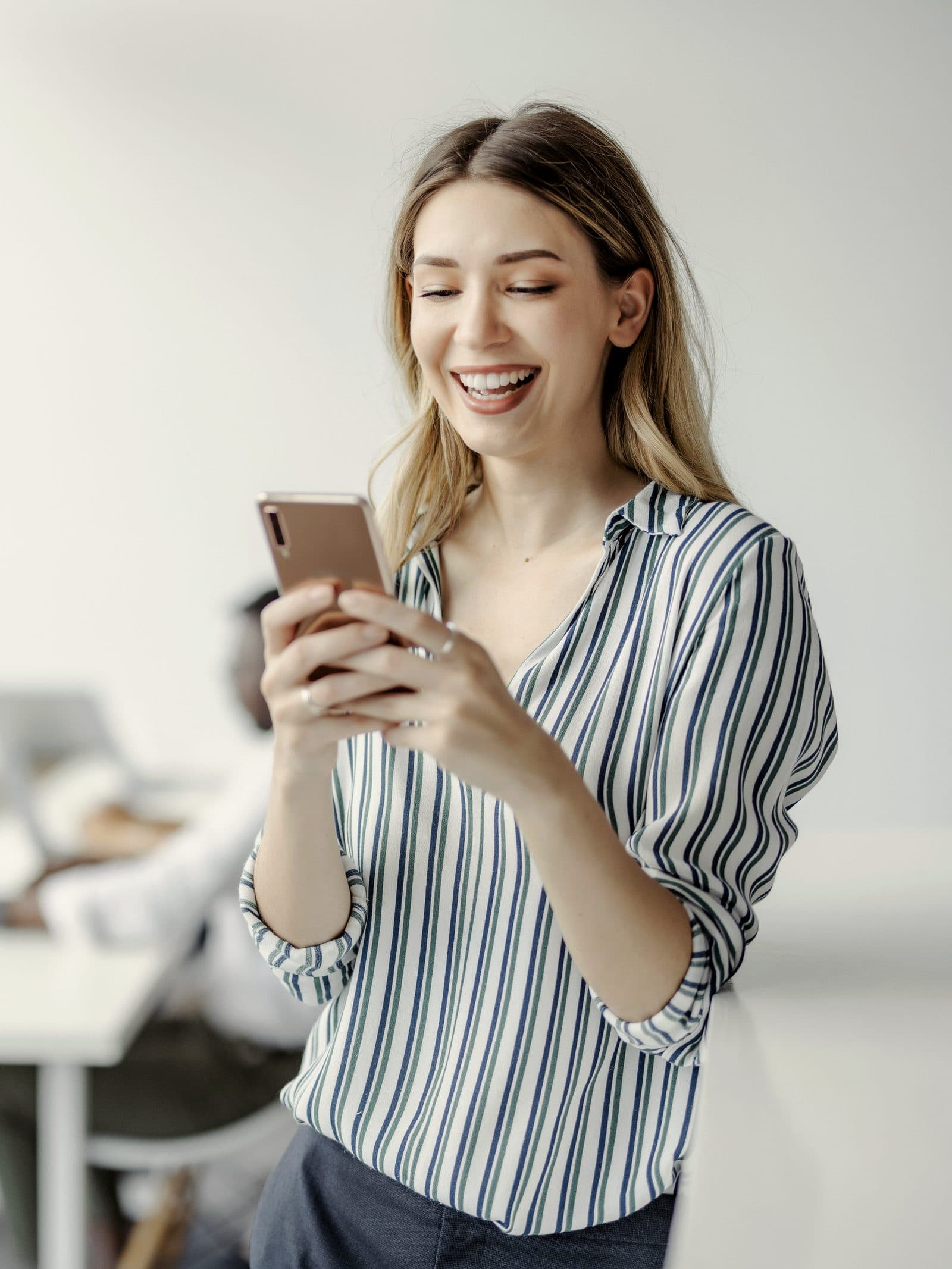 A woman smiling at an email on her phone, with colleagues working at a table behind her.